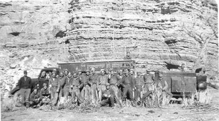A group of about 22 men posing in front of a motor vehicle that is parked by a rock wall.