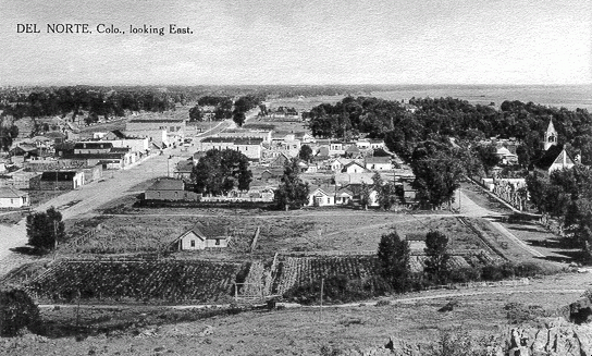 Archival photo showing a group of buildings in a neighborhood, labelled Del Norte, Colorado, looking East.